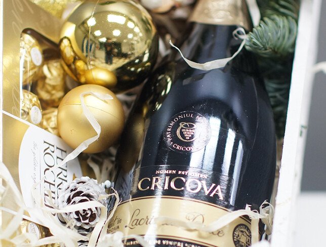Composition of a mini Christmas tree, golden baubles, Ferrero Rocher chocolates, a bottle of Cricova wine, and decorations, packed in a white wooden crate