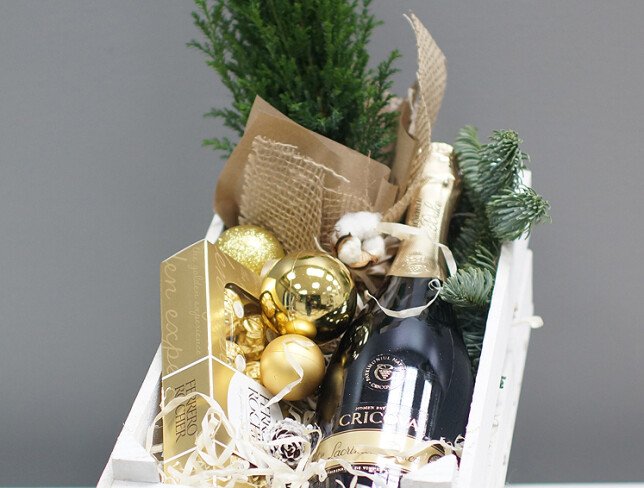 Composition of a mini Christmas tree, golden baubles, Ferrero Rocher chocolates, a bottle of Cricova wine, and decorations, packed in a white wooden crate