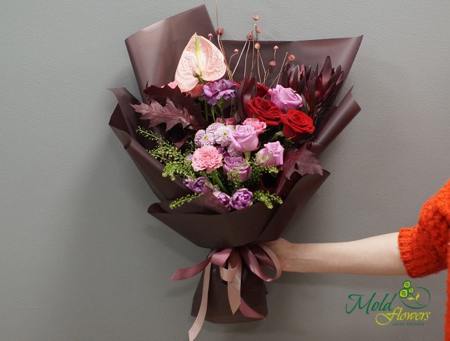 Bouquet with red and pink roses, pink anthuriums, carnations, tulips, purple chrysanthemums, scabiosa flowers, greenery, and burgundy leaves, wrapped in burgundy paper