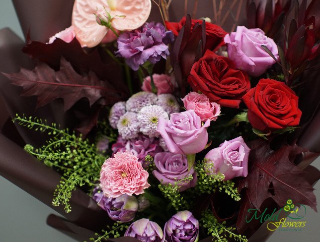 Bouquet with red and pink roses, pink anthuriums, carnations, tulips, purple chrysanthemums, scabiosa flowers, greenery, and burgundy leaves, wrapped in burgundy paper